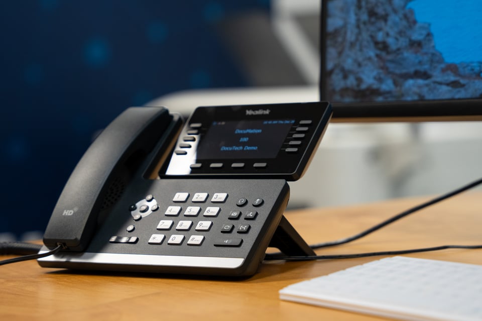 Black telephone with call routing options, dialpad, and call routing options. The brand is Yealink. In the background there is a computer screen and a white keyboard.